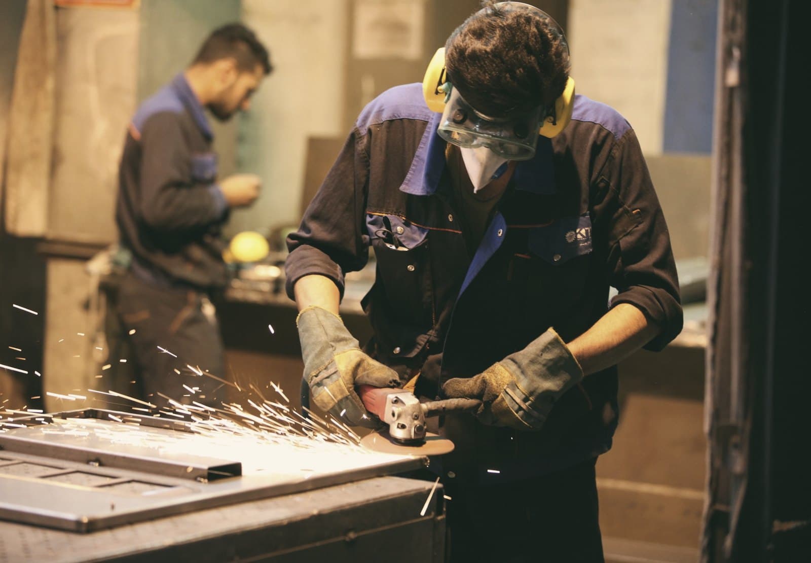 A metalworker grinding a steel part in a workshop, with a colleague behind